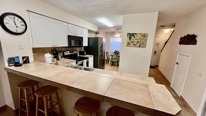 A kitchen with white cabinets, a black microwave, and a stainless steel refrigerator. There are three wooden stools at the counter. - Enjoy family time in this Gold-rated condo with quick access to the beach! (Saint Augustine)