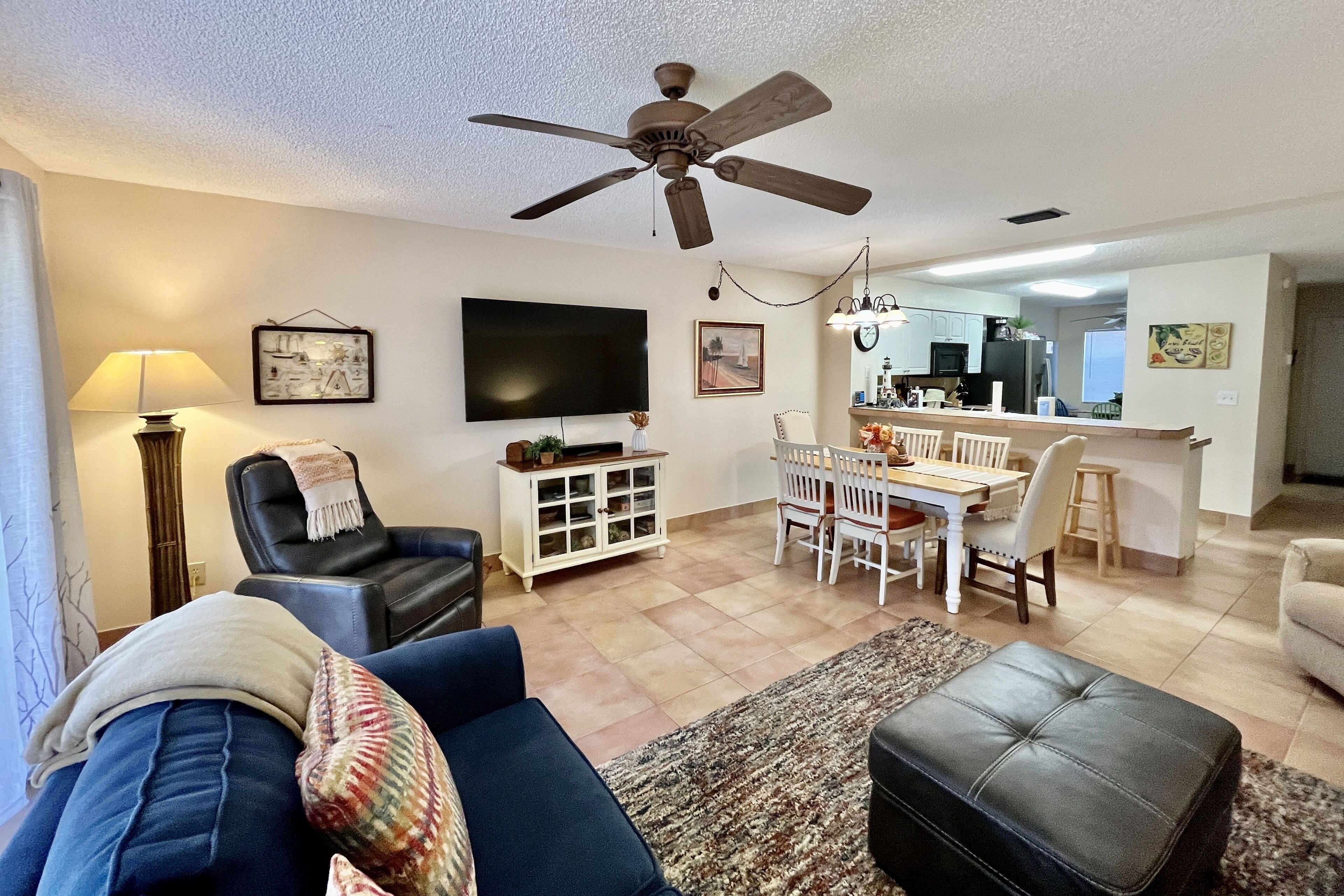 A living room with a sofa, a TV, a ceiling fan, a dining area with a table and chairs, and a kitchen in the background.