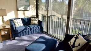 A cozy living room with a dark blue sofa, patterned cushions, and a matching ottoman. A painting hangs on the wall, and the room has large windows offering a view of palm trees. - Enjoy family time in this Gold-rated condo with quick access to the beach! (Saint Augustine)