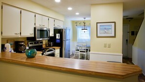 Una cocina con armarios blancos, microondas y cafetera. Hay un comedor con una mesa y sillas.