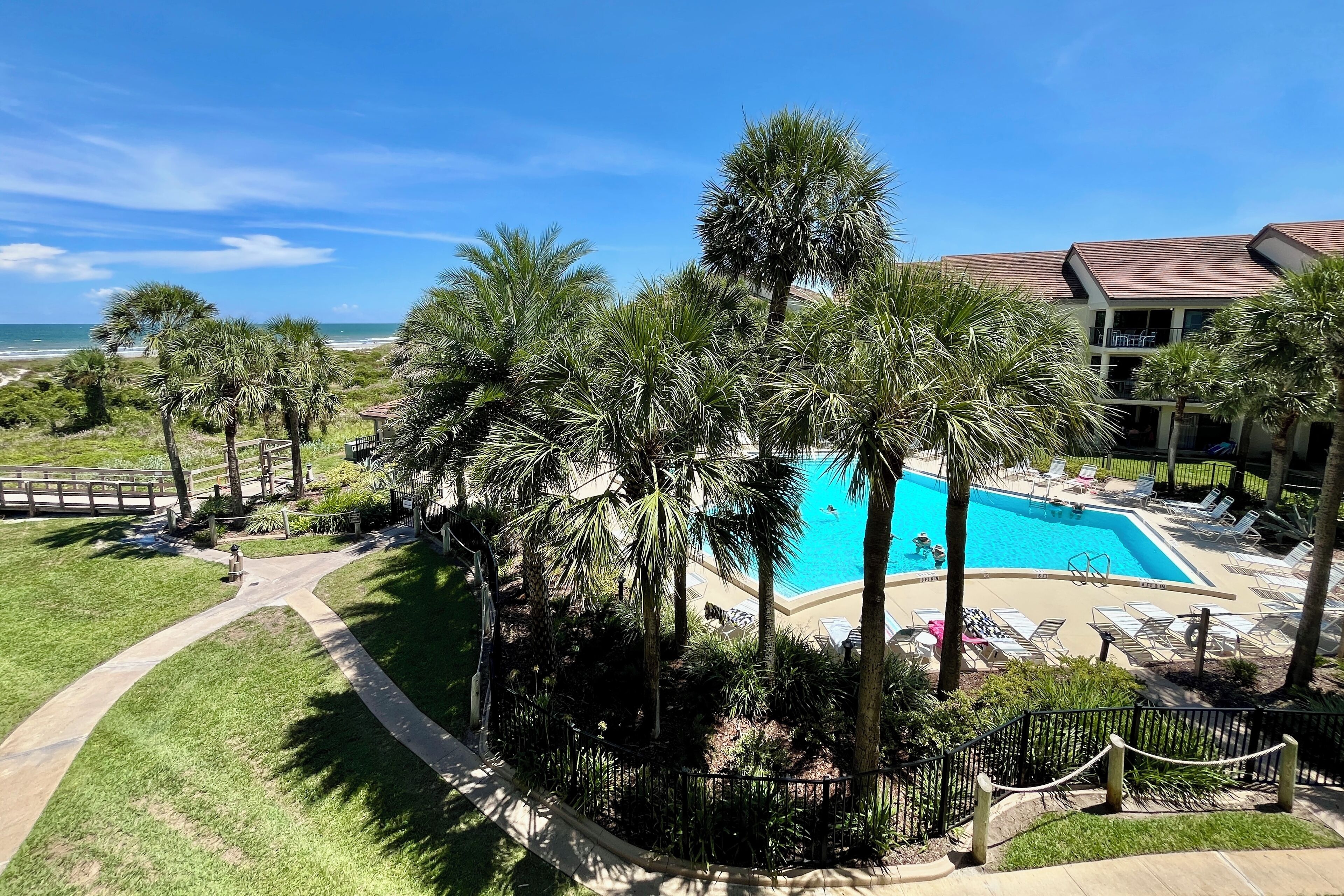 A resort with a swimming pool surrounded by palm trees, a beach view, and a building with a tiled roof.
