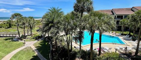 A resort with a swimming pool surrounded by palm trees, a beach view, and a building with a tiled roof.