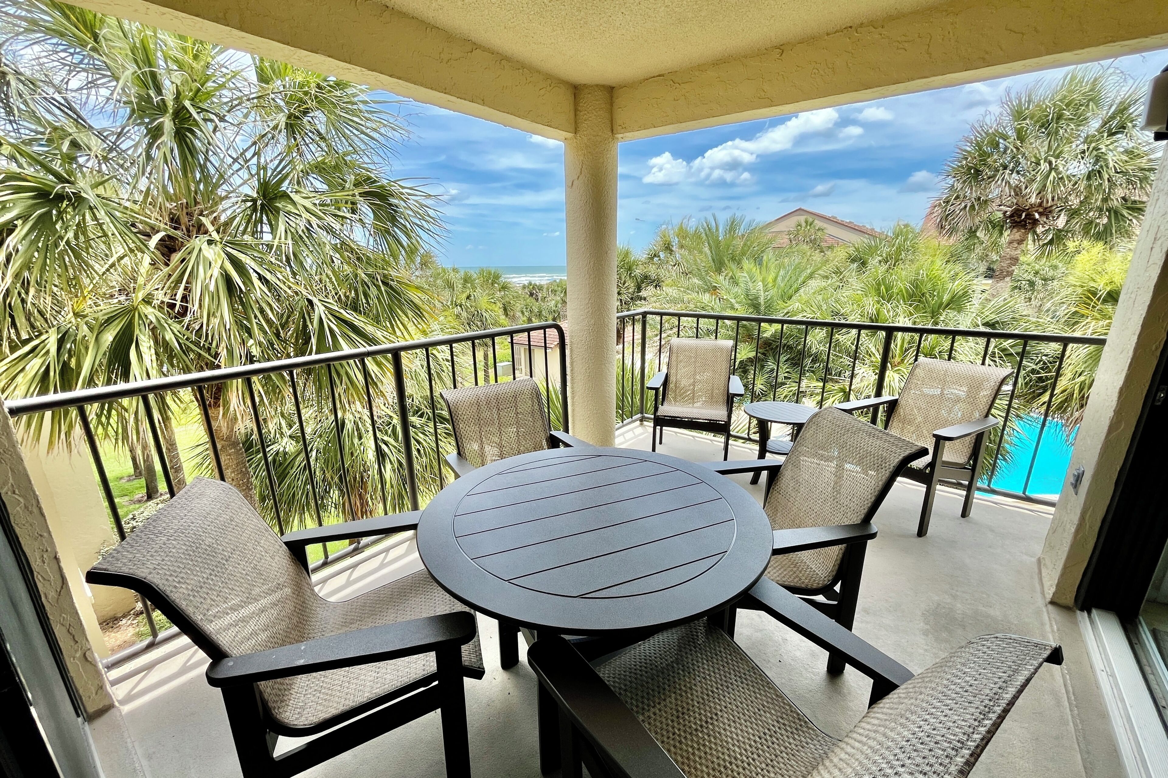 A balcony with a round table and chairs, overlooking a pool and palm trees.