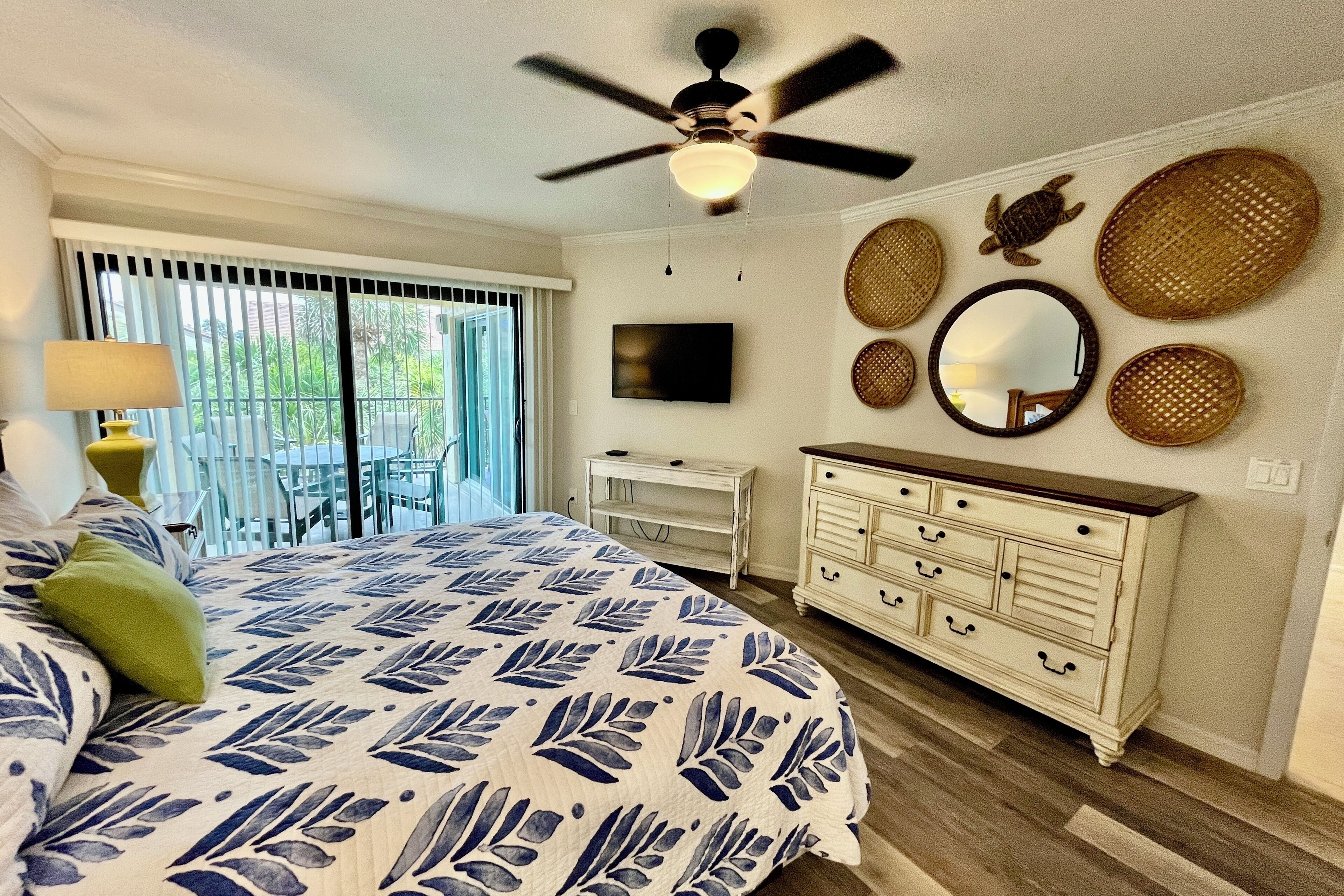 A bedroom with a bed, a dresser, a ceiling fan, a TV, and a balcony with a view of greenery.