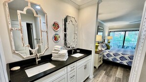 Un baño con encimera negra, muebles blancos y dos espejos grandes. En la habitación contigua, hay una cama con colcha estampada.