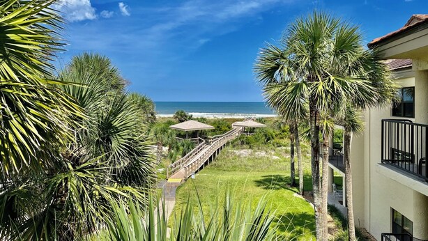 A coastal view with a wooden deck leading to beach umbrellas, surrounded by palm trees and a building with a balcony.