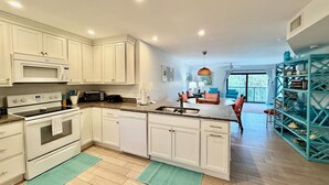 A modern kitchen with white cabinets, a built-in microwave, and a dishwasher. There is a dining area with a table and chairs, and a large window providing natural light.