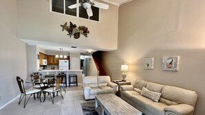 A spacious living area with a high ceiling, a dining table with chairs, a sofa, and a coffee table. There is a kitchen in the background with a staircase leading to another level. - Bright and cheery Gold-rated condo with excellent lagoon views! (Saint Augustine)