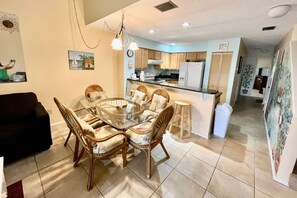 A dining area with a glass table surrounded by wicker chairs, a kitchen with wooden cabinets, and a refrigerator.