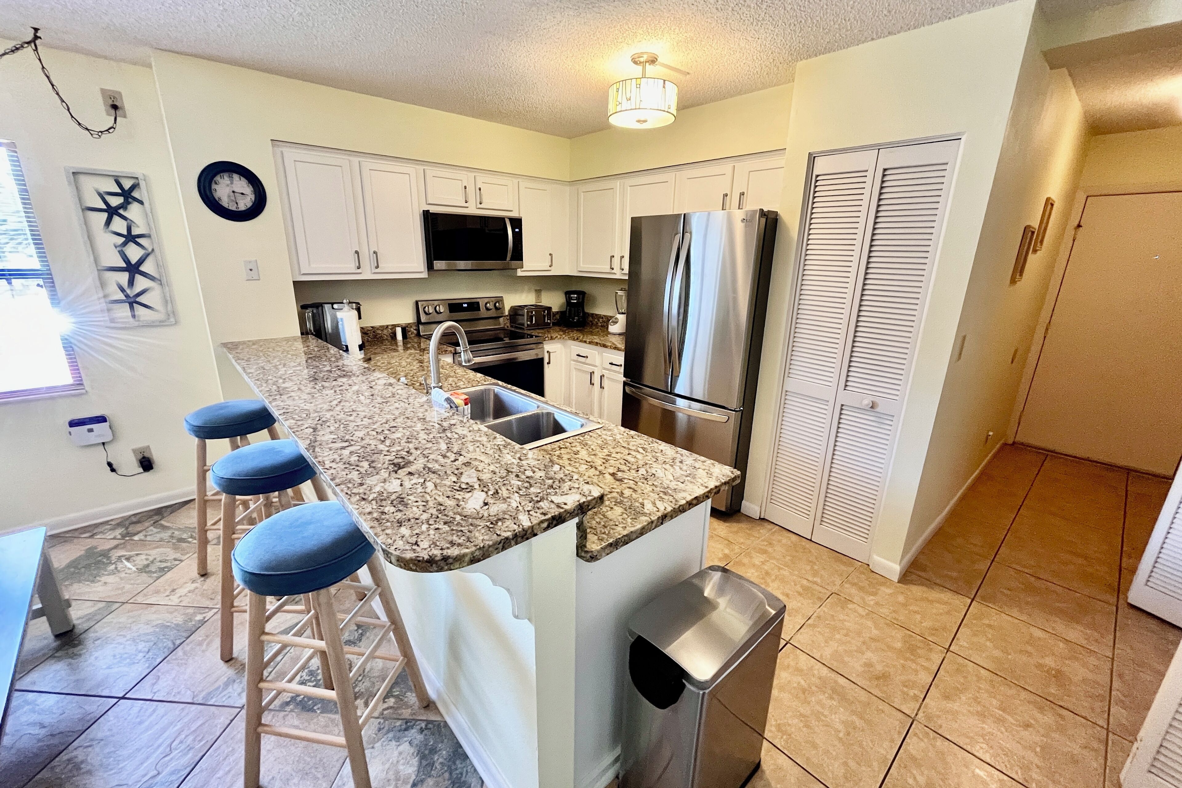 A kitchen with white cabinets, a stainless steel refrigerator, a microwave, and a sink. There are three stools and a trash can.