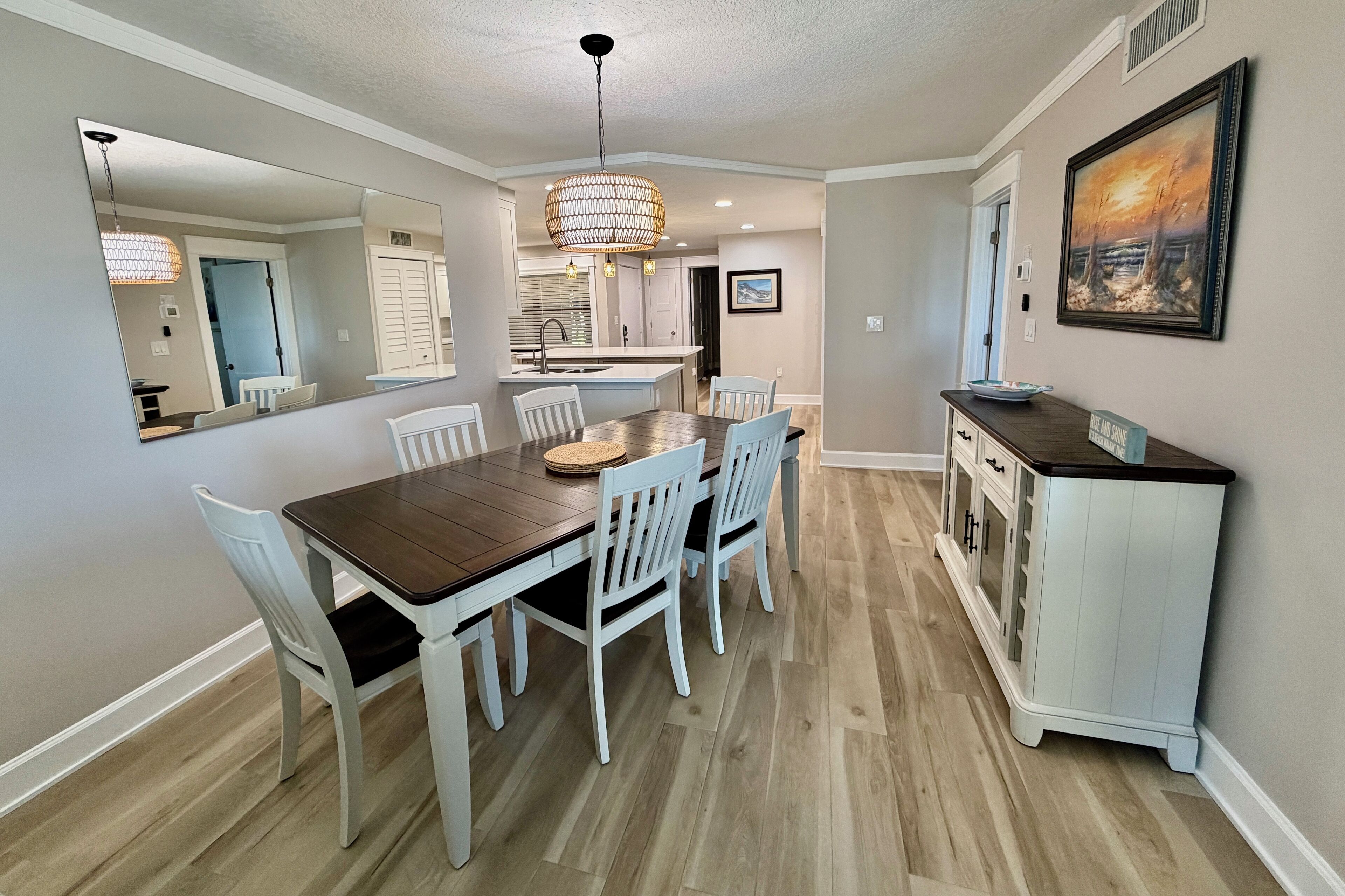 A dining area with a wooden table and white chairs, a chandelier, a kitchen in the background, and a painting on the wall.