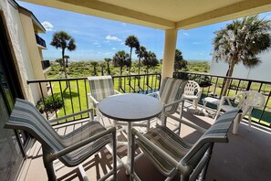 A balcony with a round table and chairs, overlooking a lawn with palm trees and a distant view. - Soak up the Florida sunshine and listen to the waves in this stunning vacation condo! (Saint Augustine)
