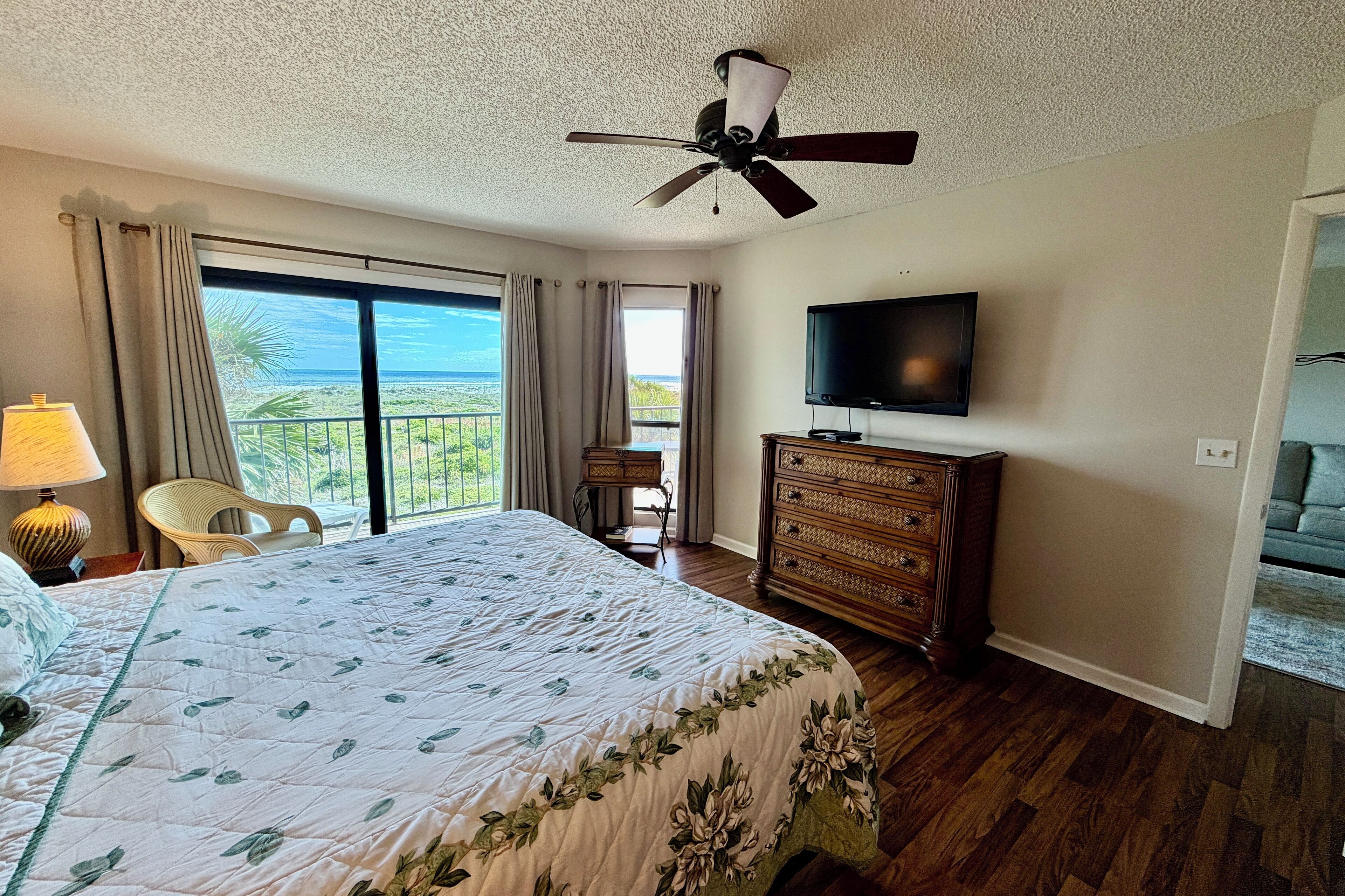 A hotel room with a large bed, a ceiling fan, a television, a dresser, and a view of the ocean through the window.