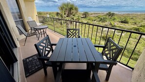 Un balcón con mesa y sillas, con vista a un paisaje costero con vegetación y cielo despejado.