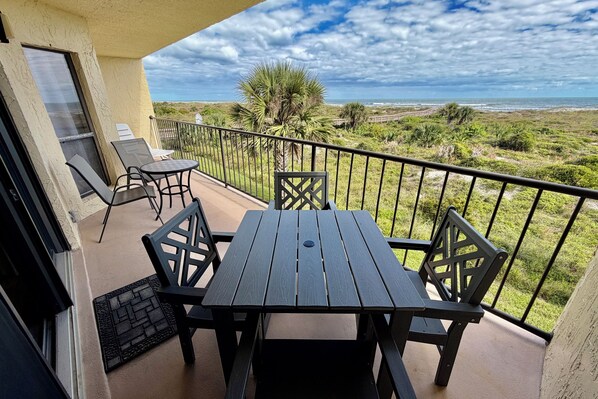 Un balcon avec une table et des chaises, donnant sur un paysage côtier verdoyant et un ciel dégagé.