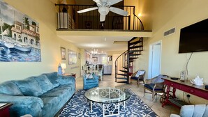 A spacious living room with a high ceiling, a ceiling fan, a large painting of a boat, a blue sofa, a glass coffee table, and a staircase leading to an upper level. - ENJOY FREE FUN EVERYDAY and a Cheerful ocean front condo with stunning view of the ocean. (Saint Augustine)