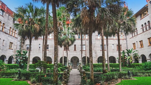 A courtyard with a central pathway, surrounded by palm trees and greenery, leading to a building with arched windows and a red roof. - Enjoy your beach vacation in this Gold-rated single family home! (Saint Augustine)