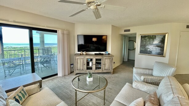 A spacious living room with a large TV, a ceiling fan, a glass coffee table, and a view of the ocean through large sliding doors. - FREE FUN DAILY with this beautiful corner unit with substantial wrap around porch and marvelous views of the Atlantic Ocean! (St. Augustine)