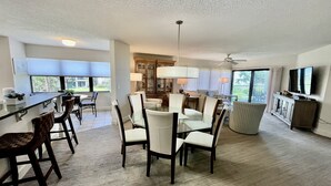 A spacious living area with a dining table, chairs, a TV, and a ceiling fan. - FREE FUN DAILY with this beautiful corner unit with substantial wrap around porch and marvelous views of the Atlantic Ocean! (St. Augustine)