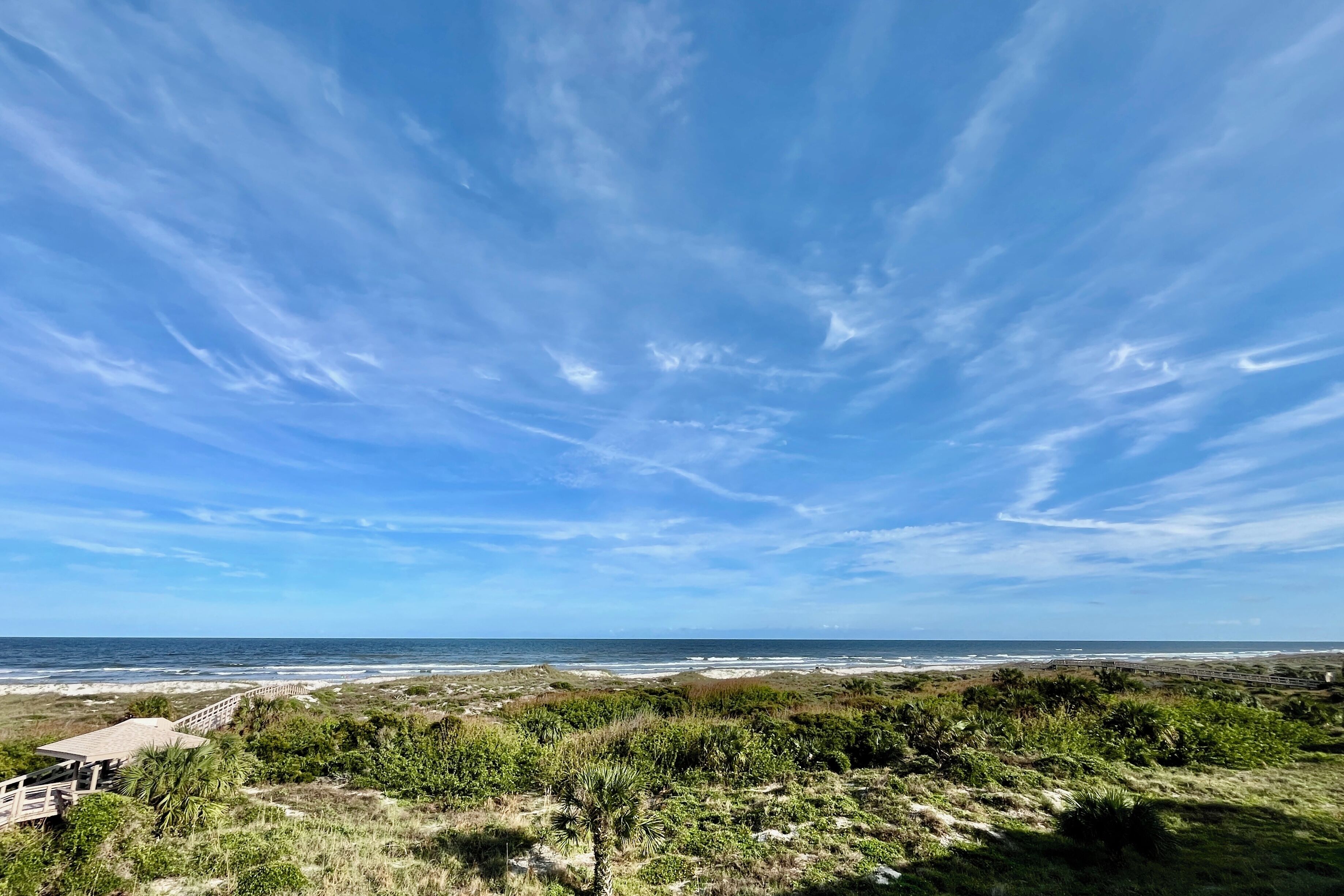 Eine Küstenlandschaft mit einem Holzsteg, der zu einem Strand führt, umgeben von Dünen und Vegetation.