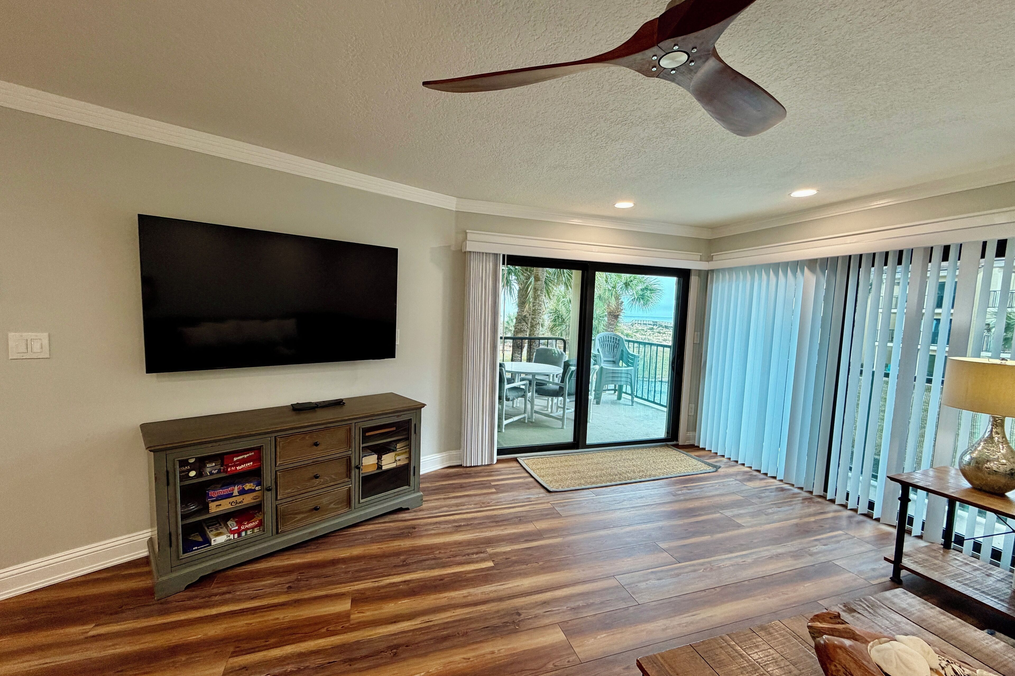A spacious living room with a flat-screen TV, wooden flooring, a ceiling fan, and a sliding glass door leading to a balcony.