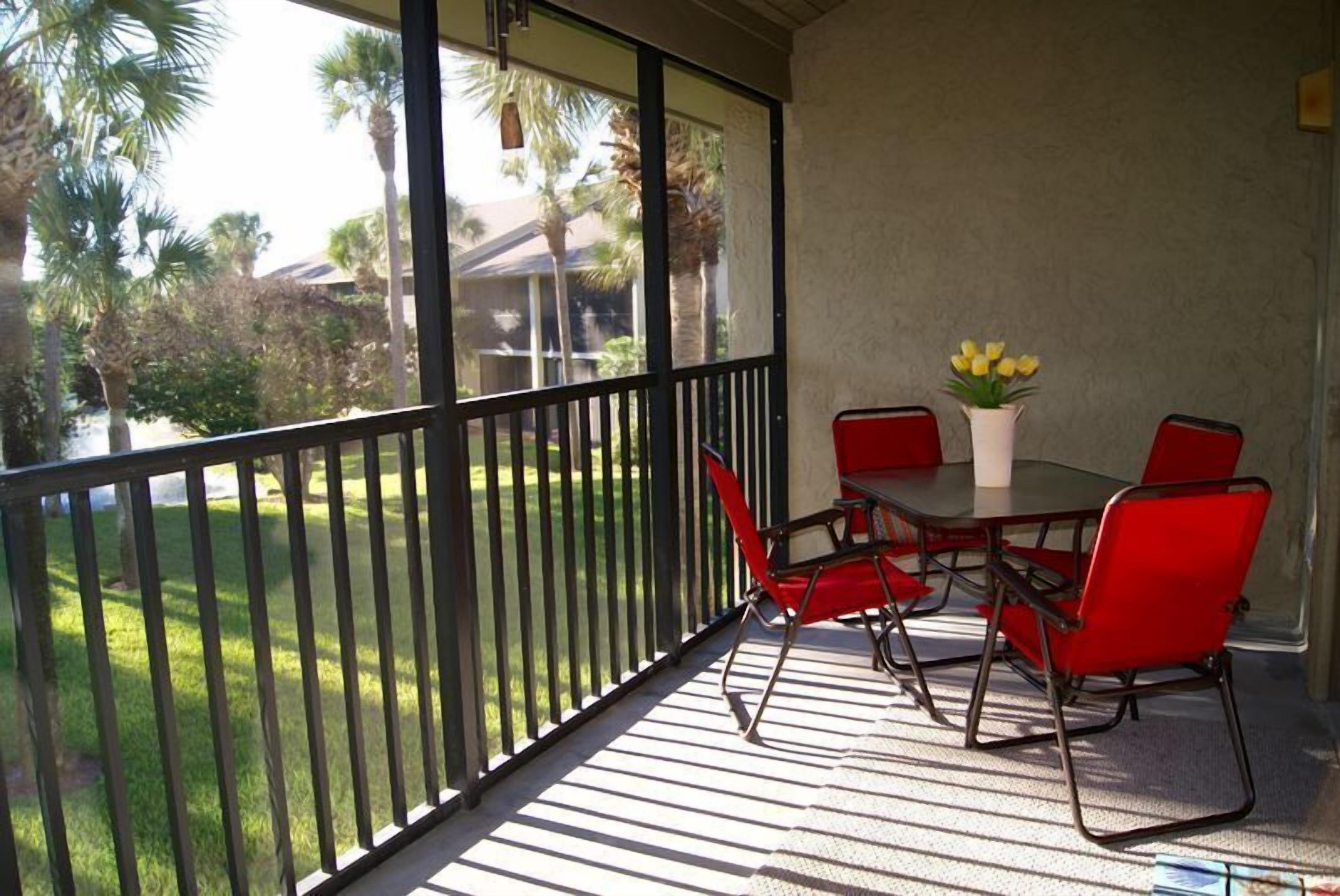 Un patio techado al aire libre con una mesa y sillas, un jarrón con flores amarillas y vistas a un césped verde y palmeras.