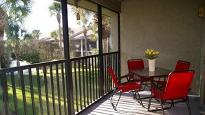 A covered outdoor patio with a table and chairs, a vase with yellow flowers, and a view of a green lawn and palm trees. - Enjoy your morning coffee on your covered lanai, then head over to beach only a brisk walk away! (Saint Augustine)