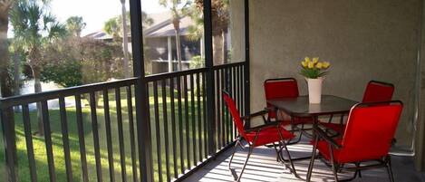 Un patio techado al aire libre con una mesa y sillas, un jarrón con flores amarillas y vistas a un césped verde y palmeras.