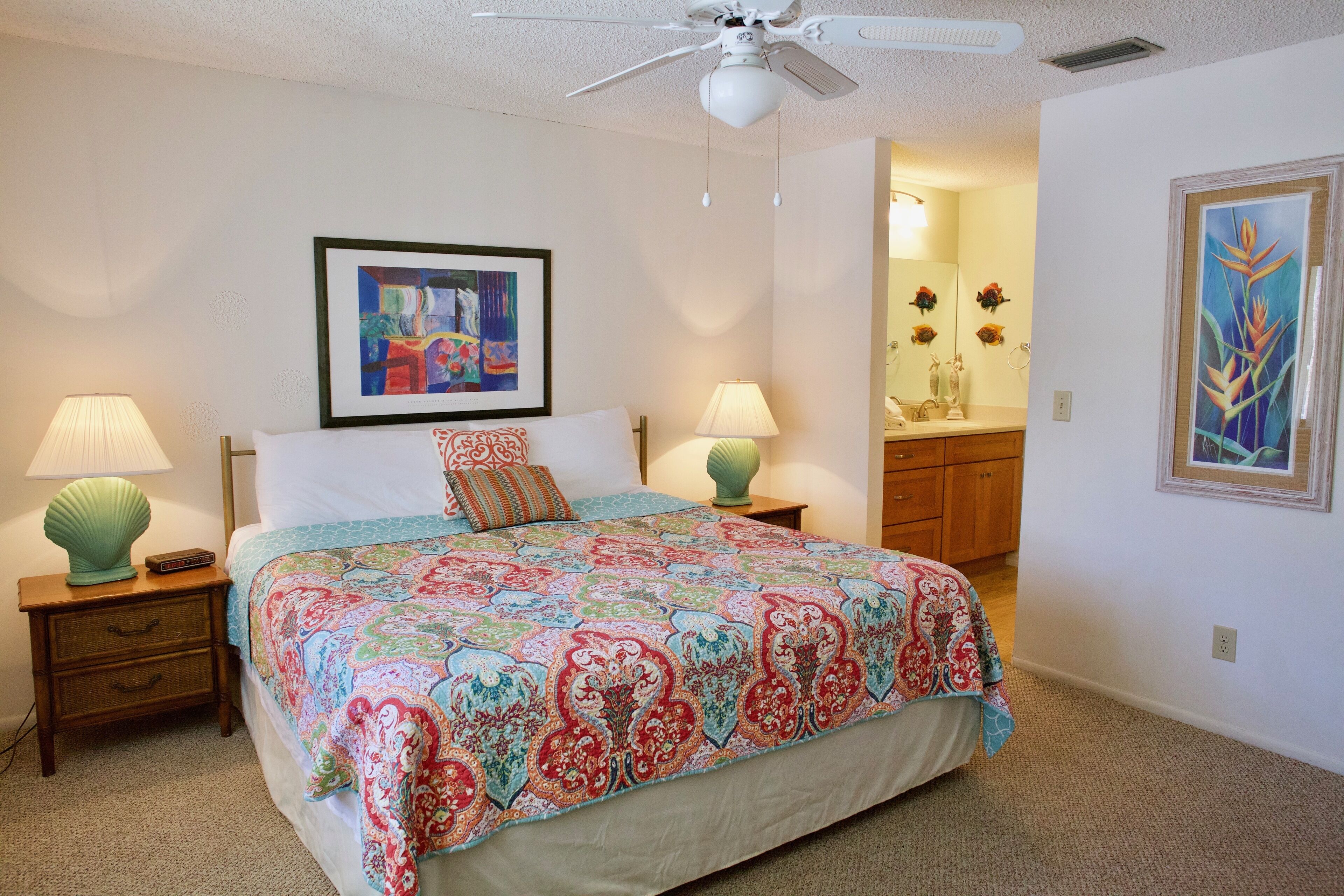 A bedroom with a large bed, two bedside tables with lamps, a ceiling fan, and artwork on the walls.