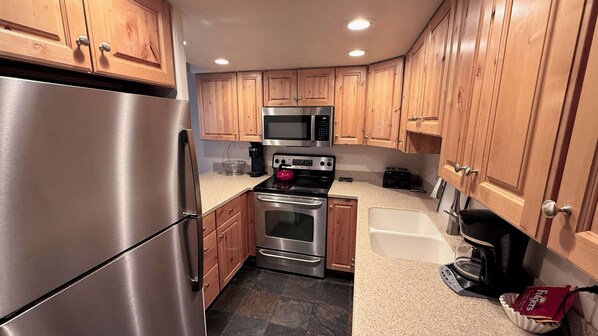 A compact kitchen with wooden cabinets, stainless steel appliances, and a stone tile floor.