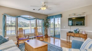 Ein geräumiges Wohnzimmer mit großem Fenster, Deckenventilator, Fernseher und Couchtisch aus Holz.
