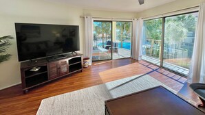 Un salon doté d’un téléviseur à écran plat, de planchers en bois, d’un ventilateur de plafond et d’une grande porte-fenêtre coulissante donnant sur un espace extérieur avec une piscine et des palmiers.