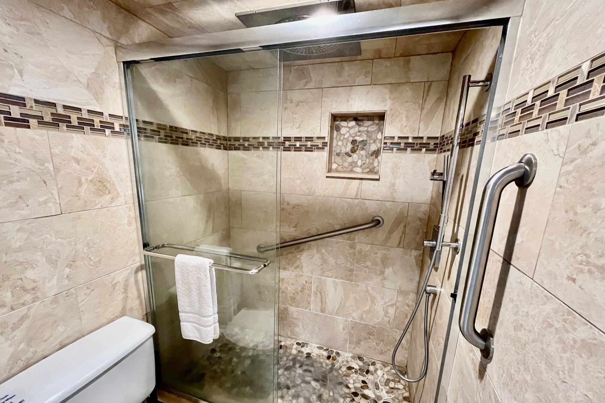 A glass-enclosed shower stall with a handrail, a small shelf, and a white towel hanging on the glass door.