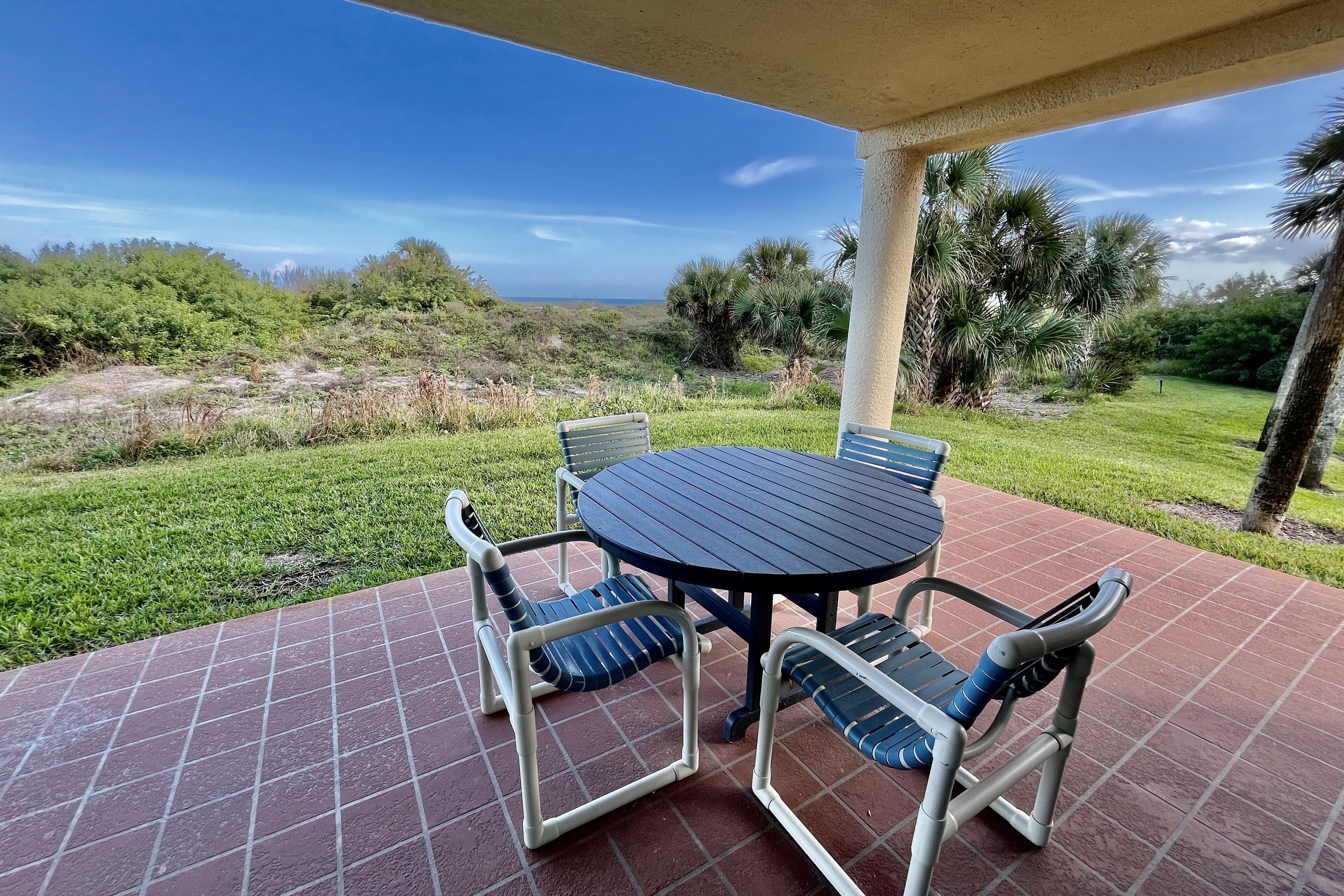 A covered patio with a round table and four chairs, overlooking a grassy area with trees and a distant view of the ocean.