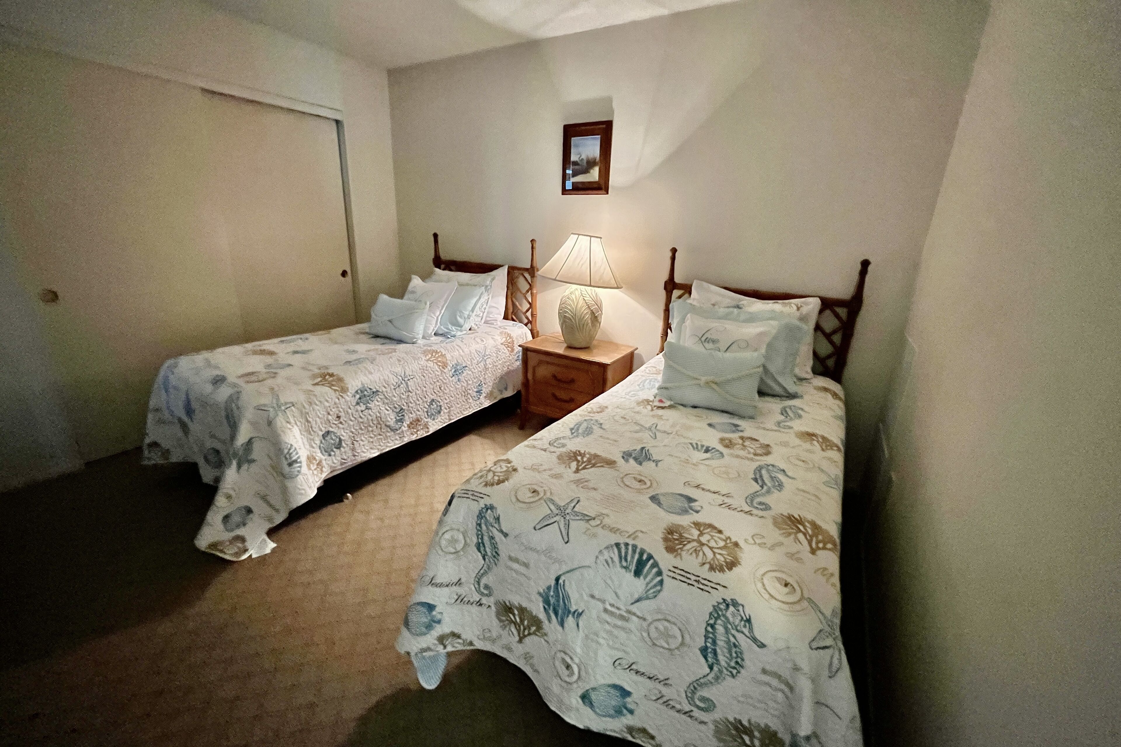 Une chambre avec deux lits simples, une table de chevet avec une lampe et un tableau encadré au mur.