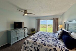 A bedroom with a bed, a television on a dresser, a chair, and a view of the outdoors.