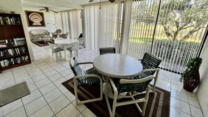 Une terrasse ensoleillée avec une table ronde et des chaises, un ventilateur de plafond, une bibliothèque et une vue sur un jardin, visible à travers de grandes fenêtres.