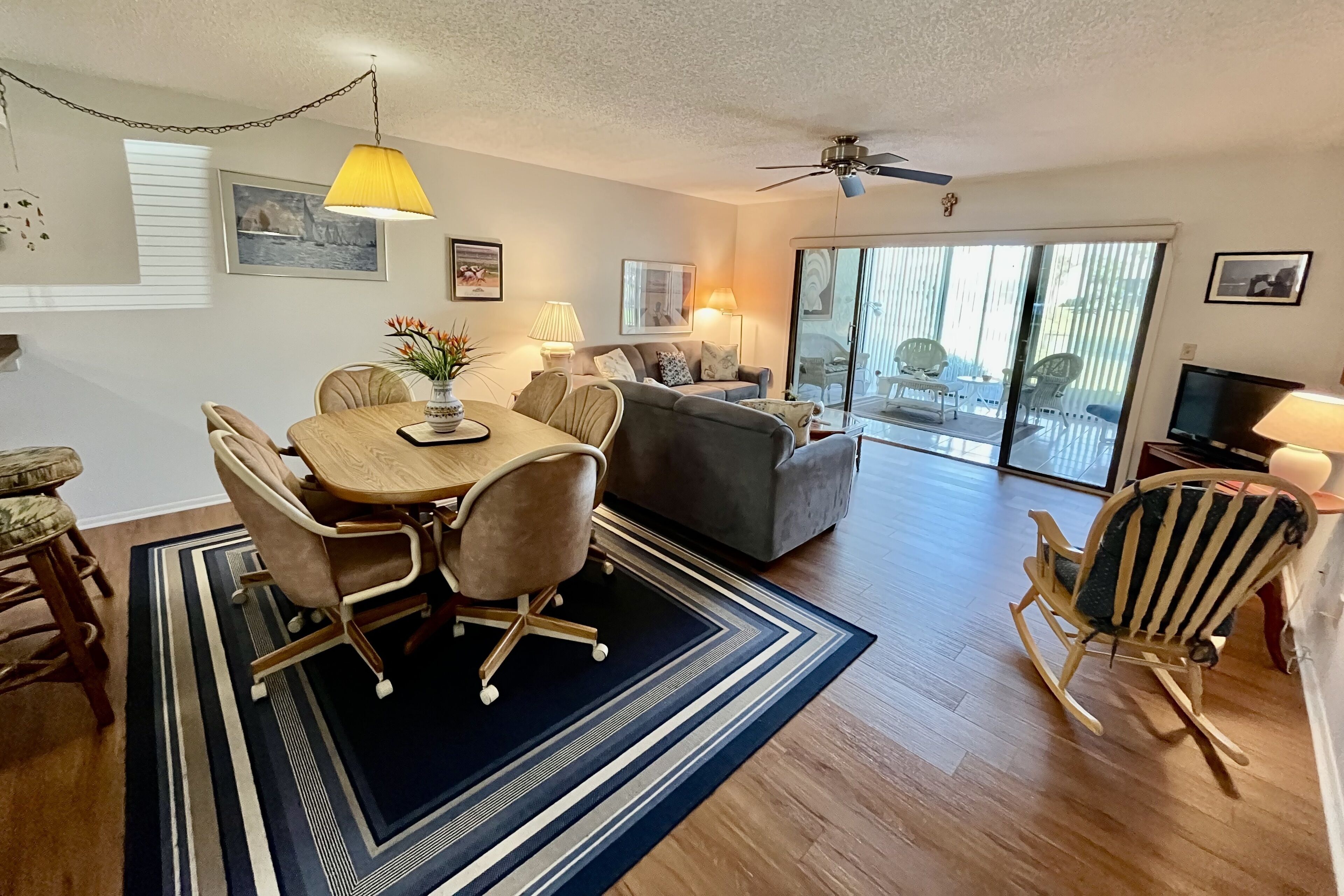 A spacious living area with a dining table, chairs, a sofa, and a rocking chair. There is a ceiling fan, a TV, and a large window with a view of the outdoors.