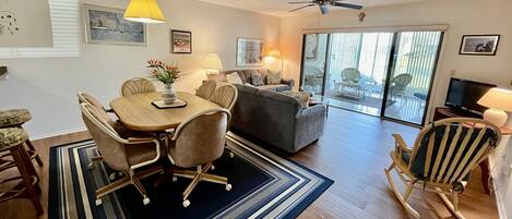 A spacious living area with a dining table, chairs, a sofa, and a rocking chair. There is a ceiling fan, a TV, and a large window with a view of the outdoors.