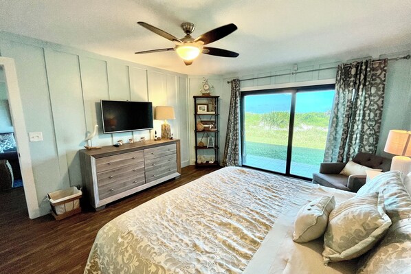 Ein Schlafzimmer mit einem großen Bett, einem Deckenventilator, einem Fernseher, einer Kommode und Blick ins Freie durch eine Glastür.