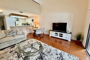 A living room with a sofa, a glass coffee table with decorative items, a flat-screen TV on a white cabinet, and a dining area with chairs and a table.