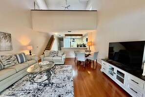 A spacious living room with a sofa, coffee table, and TV. There is a staircase leading to an upper level with a kitchen.