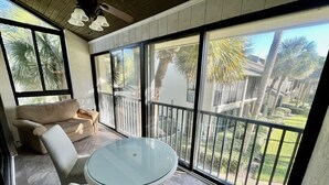 A sunlit room with a glass table and chairs, a beige sofa, and large windows with a view of a backyard and palm trees. - Comfortable condo with easy beach and pool access! (Saint Augustine)