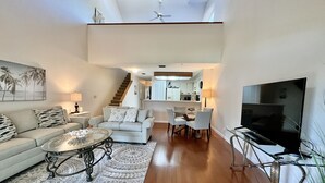 A modern living room with a sofa, coffee table, and TV. There is a staircase leading to a kitchen and dining area. - Comfortable condo with easy beach and pool access! (Saint Augustine)