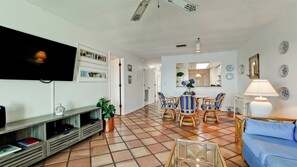 A living room with a TV, a dining area with striped chairs, a blue sofa, and a tiled floor. - Gulf Watch 216 - Anna Maria Island - Condo 2 Bedroom / 2 Bath Gulf to Bay access, maximum occupancy of 4 people . (Bradenton Beach)