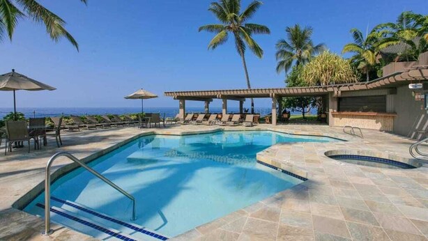 A pool area with lounge chairs and umbrellas, palm trees, and a view of the ocean. - Lush View in Sunny Kona-Keauhou! Kitchen Ease, Lanai, Laundry, WiFi, TV Kanaloa 1204 (KAILUA-KONA)