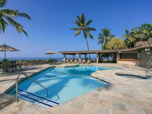 A swimming pool with lounge chairs and umbrellas, surrounded by palm trees and a clear view of the ocean.