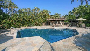 Una piscina con suelo de baldosas, rodeada de palmeras y una zona de estar cubierta.