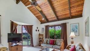 Un salón con techo de madera, un televisor sobre un mueble, un sofá, una mesa de centro y una ventana con cortinas.