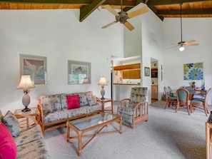 A spacious living room with a sofa, chairs, a coffee table, and a dining area with chairs and a table. There are ceiling fans, wall art, and a kitchen area visible.